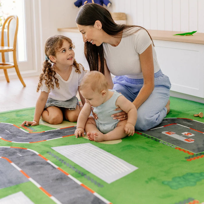 A woman and two young girls sit on a green play rug with road and track graphics in a bright indoor room. The product being shown is the printed play rug rather than the children's clothing.