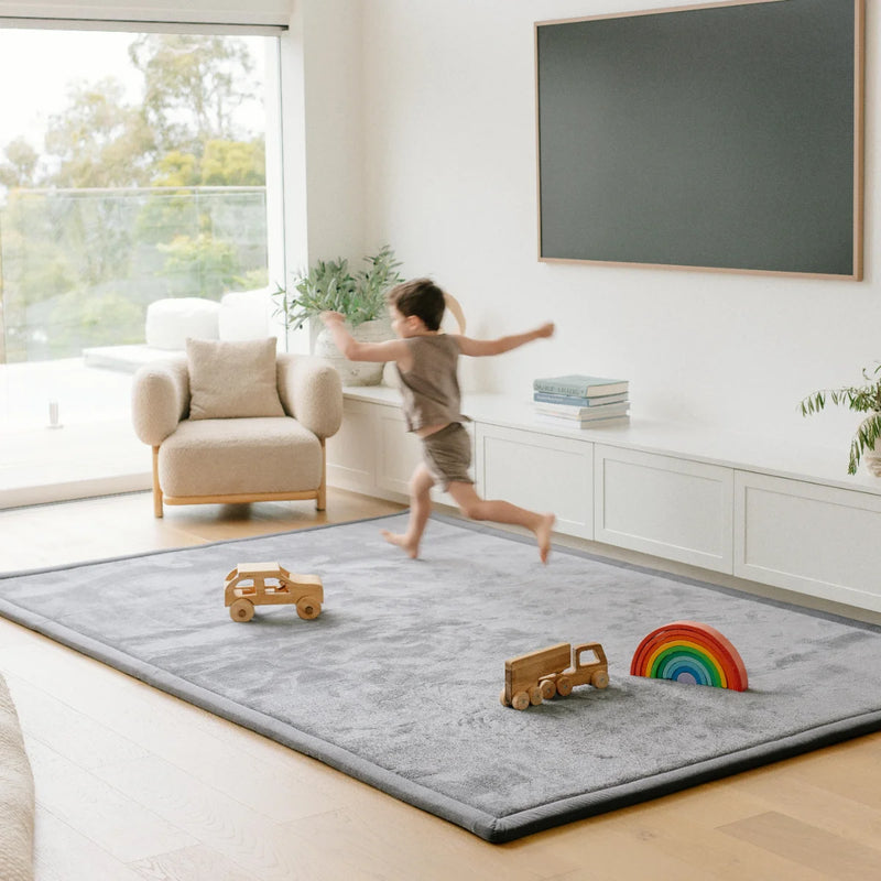 A child runs across a large gray plush play rug in a bright, modern living room. The rug has a black piped edge and is styled with wooden toy trucks and a rainbow toy.